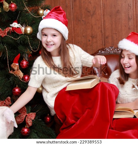 Indoor portrait of happy smiley little girls playing with lovely dog on Christmas time/Beautiful little girls wearing Santa hats holding books celebrating Christmas holiday
