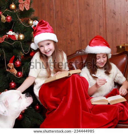 Indoor portrait of happy smiley little girls playing with lovely dog on Christmas time/Beautiful little girls wearing Santa hats holding books celebrating Christmas holiday