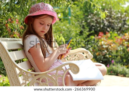 Cute smiling little girl with enthusiasm draws in album sitting in the summer park bench
