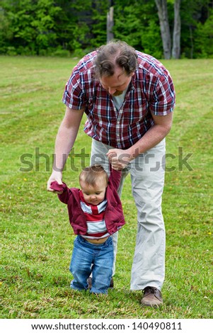 Father helping toddler son to learn how to walk in an outdoor setting
