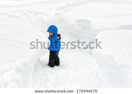 A small toddler boy points the way through pathways carved in snow accumulation taller than he is. Typical winter weather within the lake effect or snow belt regions of United States and Canada.