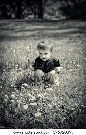 A young boy sits in a field holding a dandelion flower.  Black and white image toned for a vintage look.
