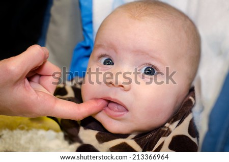 An adult\'s finger is shown checking inside a baby\'s mouth. The teething baby has a funny expression.