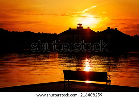 A silhouetted park bench on a dock by the lake.  Beyond the lake the sun sets behind a building.