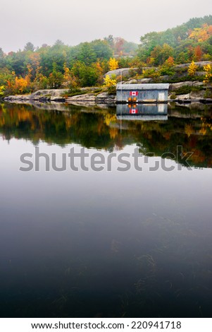 An old wooden boat house with a Canada flag on its wall is on a calm lake surrounded by a rocky landscape and colorful trees on a misty autumn morning.