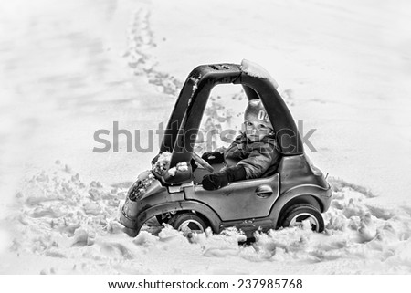 A young boy dressed for cold weather sits in a red toy car stuck in the snow during the winter season.  Processed in black and white.  Filtered for a retro, vintage look.