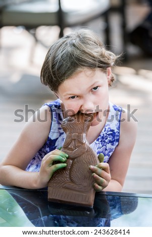 A girl sits outside takes a bite out of a large chocolate bunny on Easter day during the spring season.