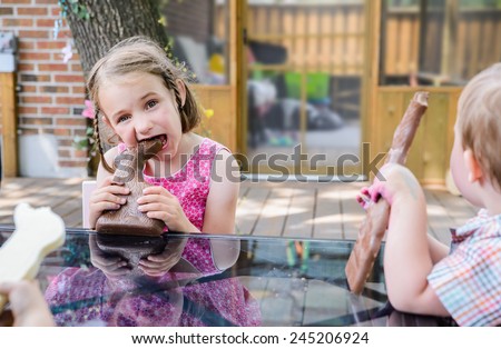 A little girl in a pink dress sits outside taking a big bite out of a large chocolate bunny on Easter day during the spring season.  Part of a series.