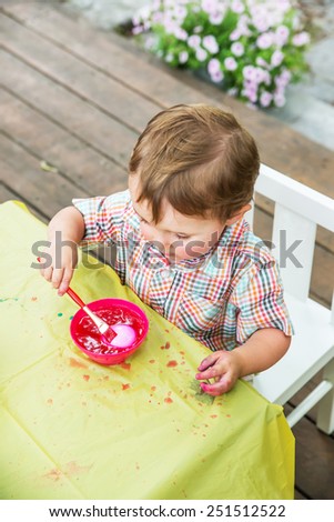 A happy boy sitting outside in the garden during the springtime having fun painting and decorating Easter eggs.  He\'s paint brushing his egg in a bowl with pink color dye.  Part of a series.