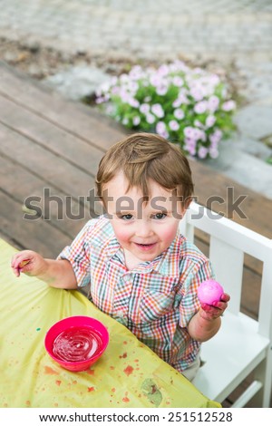 A happy boy sitting outside in a springtime garden setting at a crafts table painting and decorating Easter eggs.  He smiles proudly and holds up a freshly colored pink dyed egg.   Part of a series.