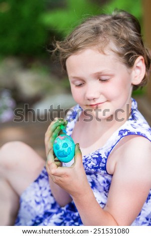 Happy little girl in a pretty dress smiles and decorates a color dyed Easter egg with paint while outdoors in the garden setting during the springtime.  Selective focus on the Egg.  Part of a series.