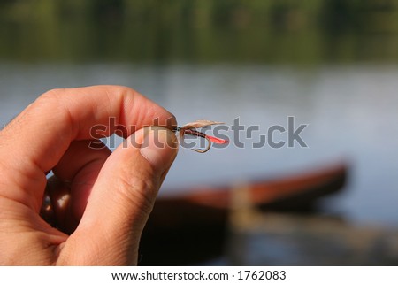 Fly fisherman holds up a colorful hand tied fly with lake and canoe in the background