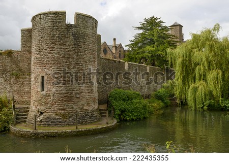 round tower and willow tree on moat at Bishop palace ,Wells view of tower and ancient walls on moat of historic palace,  shot in bright light under a cloudy sky with a big willow tree nearby