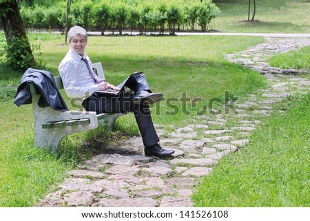 A young handsome businessman using laptop sitting on a bench in a park./Businessman using laptop sitting on a bench