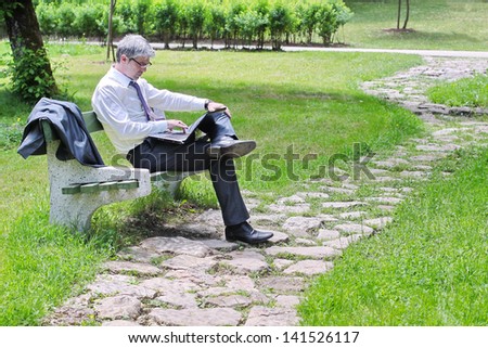 A young handsome businessman using laptop sitting on a bench in a park... looking and smiling into camera./Businessman using laptop sitting on a bench