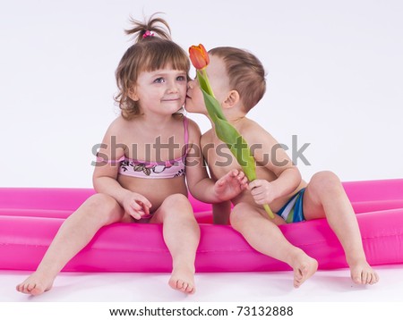 Two gay children in swimsuits sitting on a big air mattress.A boy kisses a girl and want to give her flowers.