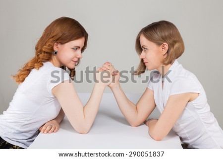 two pretty ladies in white blouses arm wrestling. female rivalry, friendship, dispute, competition. business concept. portrait on grey background.