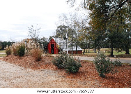 Old red barn guarding with much dignity over the pasture that lies beyond.  Trees, shrubs and a road elegantly enhance this barn with a white birdfeeder in the foreground.
