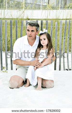Beautiful little girl sitting on her daddy\'s knee by a wooden fence in the sand along the shore.