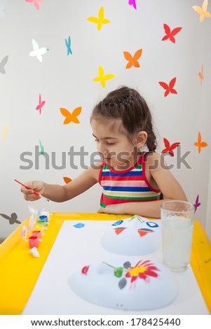 Beautiful girl in bright clothes sitting at the table and painting. Colorful false butterflies on background.