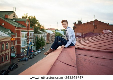 Young bold guy girl woman in hipster clothes, sit on edge of roof, dressed like a boy man in shirt, bow-tie, suspenders and pants trousers on background of city, road and jam