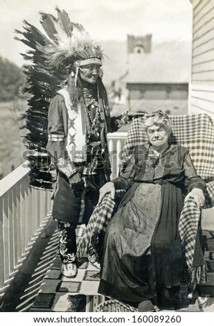 US - Wyoming - CIRCA 1900 A vintage photo of an Indian Chief in full headdress standing next to an older elderly woman sitting in a chair. CIRCA 1900