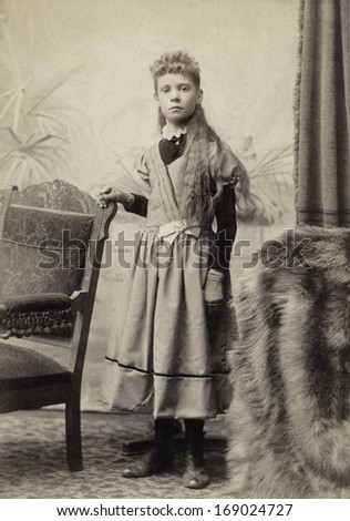 USA - OHIO - CIRCA 1898 - A vintage antique photo of a young girl dressed in a Victorian style dress with long hair. She is standing next to a chair. A photo from the Victorian era. CIRCA 1898