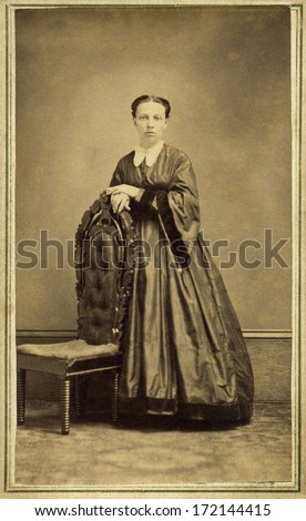 USA - WISCONSIN - CIRCA 1865  A vintage Cartes de visite photo of young pioneer woman standing chair. She is dressed in hoop skirt dress with pagoda sleeves. A photo from the Civil War era. CIRCA 1865