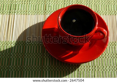 Red coffee cup and saucer filled with coffee sitting on a place mat. Sunlight comes in from upper right corner producing a shadow to the left.