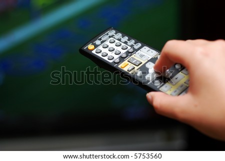 A hand holding a TV remote control and pointing it toward a television screen showing a football game. Selective focus. TV image is out of focus.