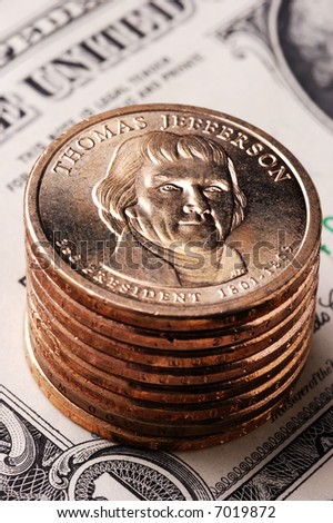 Vertical close-Up of a Jefferson dollar coin on a stack of dollar coins with an out-of-focus paper dollar bill in the background.