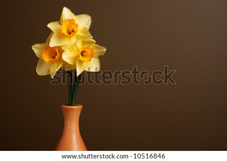 Three daffodils in a simple wooden vase on a brown background with space for text on the right side.