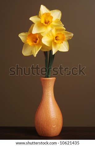 Vertical image of three daffodils in a simple wooden vase with a brown background.