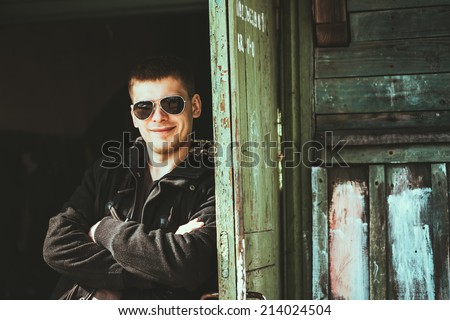 Young handsome man staying near old wooden house in autumn day, relaxing,  thinking, smiling. Casual style - jacket, sunglasses.