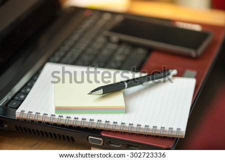 Opened notepad with pen, small sticky paper and black smart phone on laptop computer in coffee shop with morning scene