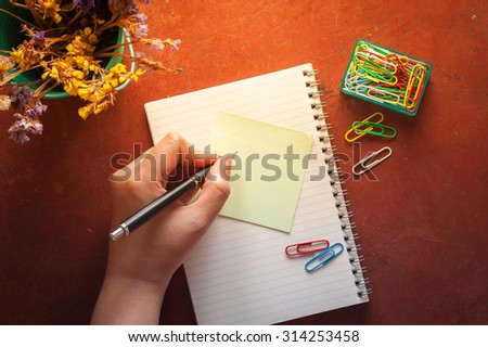 Opened notepad with pen, small sticky paper, and paper clips with female left hand writing on blank area on red cement background with morning scene
