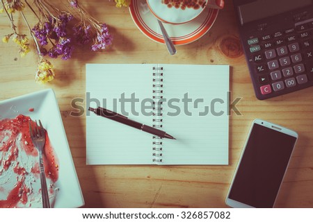 Opened notebook with blank area for text or message, pen, smart phone, dirty dessert dish, and calculator on wood table in afternoon time with film filter effect