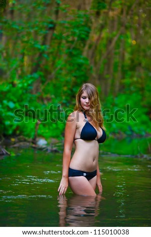 Blonde nude young woman big breasts in a black bikini standing on a river in the woods with his hands behind his head