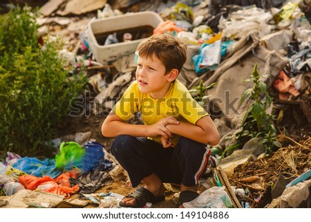 German homeless baby boy squatting on rubbish a dump in yellow t-shirt with tousled hair