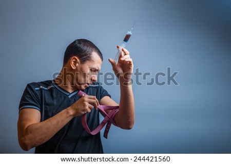 male of European appearance brunet harness tightens and holds a syringe in his hand, a drug addict on a gray background