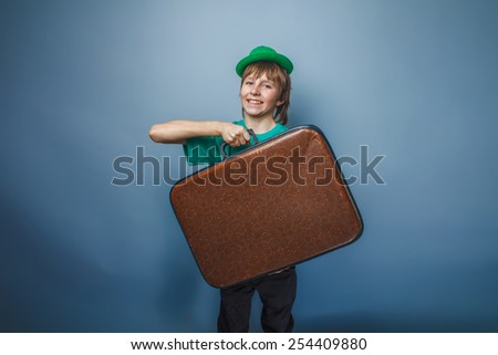 European appearance teenager boy in a shirt in green hat holds an old suitcase in hand on gray background, adventure