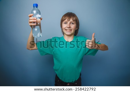 boy teenager European appearance in a green t-shirt showing sign okey holding a bottle of water on a gray background