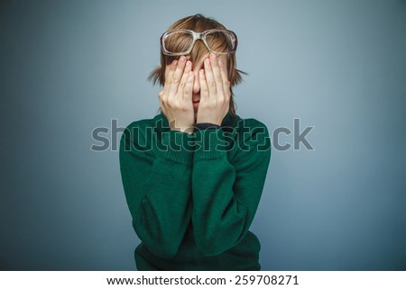 European-looking boy of ten years in glasses covered his face with his hands on a gray background