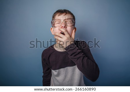 teenager boy of European appearance with glasses covers her mouth yawns on a gray background, drowsiness, fatigue