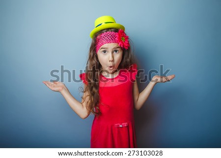 Girl European appearance haired child of seven years in a bright dress in yellow hat surprised on a gray background, spread her hands