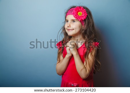 Girl European appearance haired child of seven in red bright dress praying on a gray background, smile
