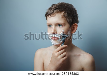 Teen boy shirtless European appearance in brown hair foam on face shaving razor safe on a gray background, concentration, seriousness, growing