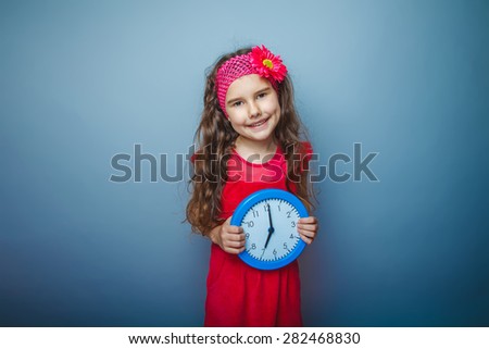 a girl of seven European appearance brunette in a bright dress holding a blue watch on gray background, time, smile