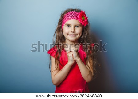 a girl of seven European appearance brunette in a bright dress folded her hands in prayer, watching in front of a gray background, smiling, portrait