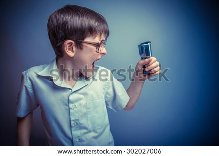 A boy of about ten European appearance in light brown shirt and glasses holding a camera action and shouts against the gray background, videography, fury retro photo effect